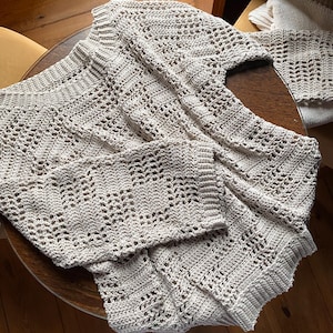 Pu&ograve; includere: Un maglione all'uncinetto beige chiaro a trama aperta con maniche lunghe, appoggiato su un tavolo di legno. Il maglione ha uno scollo a V e un design strutturato e fantasia. Un pezzo all'uncinetto abbinato &egrave; visibile sullo sfondo.
