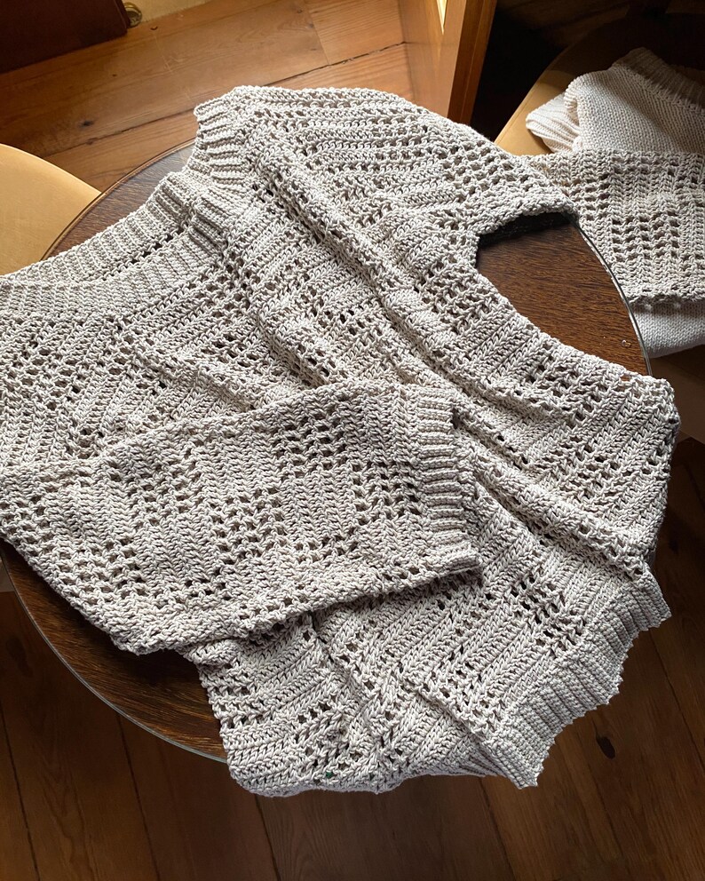 Pu&ograve; includere: Un maglione all'uncinetto beige chiaro a trama aperta con maniche lunghe, appoggiato su un tavolo di legno. Il maglione ha uno scollo a V e un design strutturato e fantasia. Un pezzo all'uncinetto abbinato &egrave; visibile sullo sfondo.