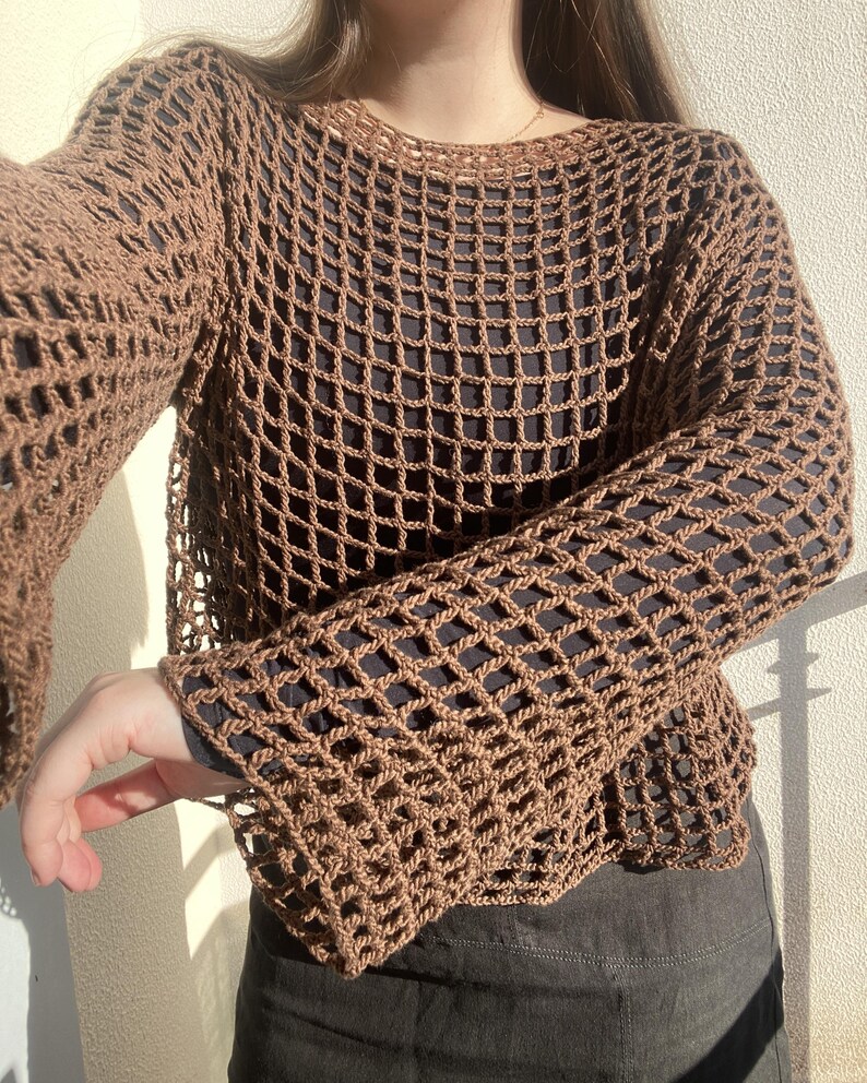 May include: A brown crochet top with long sleeves and a net-like pattern. The open weave design is visible. The top is worn over a black garment, creating a contrast. The sleeves are flared at the wrist.