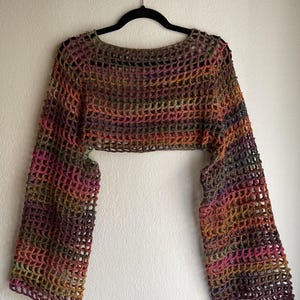 May include: A multi-coloured crochet shrug with long sleeves, hanging on a black hanger. The open-weave design features a blend of colours including brown, pink, orange, and green. The garment is a cropped style, suitable for layering.