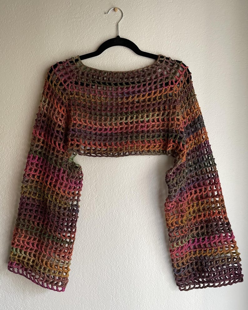 May include: A multi-coloured crochet shrug with long sleeves, hanging on a black hanger. The open-weave design features a blend of colours including brown, pink, orange, and green. The garment is a cropped style, suitable for layering.