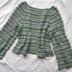 May include: A green, grey, and white crochet top with long sleeves. The top has a scoop neckline and a flared hem. The crochet pattern includes horizontal stripes and openwork details, creating a textured appearance. The sleeves are bell-shaped.