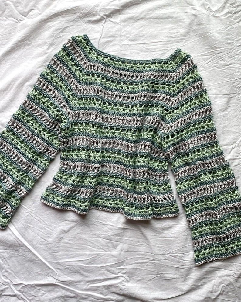 May include: A green, grey, and white crochet top with long sleeves. The top has a scoop neckline and a flared hem. The crochet pattern includes horizontal stripes and openwork details, creating a textured appearance. The sleeves are bell-shaped.
