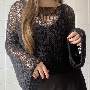 May include: Black open-weave knit top with long, flared sleeves. The top has a scoop neckline and is worn over a black camisole. The fabric appears to be a loosely woven yarn, creating a net-like effect. The sleeves are wide and flowing.