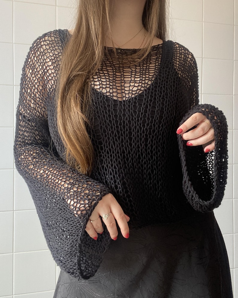 May include: Black open-weave knit top with long, flared sleeves. The top has a scoop neckline and is worn over a black camisole. The fabric appears to be a loosely woven yarn, creating a net-like effect. The sleeves are wide and flowing.