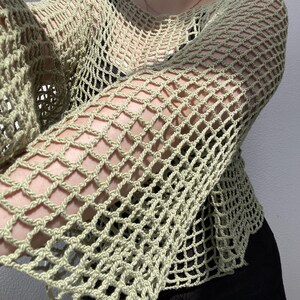 May include: A pale green, open-weave crochet top with long sleeves. The top has a loose, net-like pattern, creating a see-through effect. The garment is designed with a round neckline and a relaxed fit, suitable for layering or as a standalone piece.