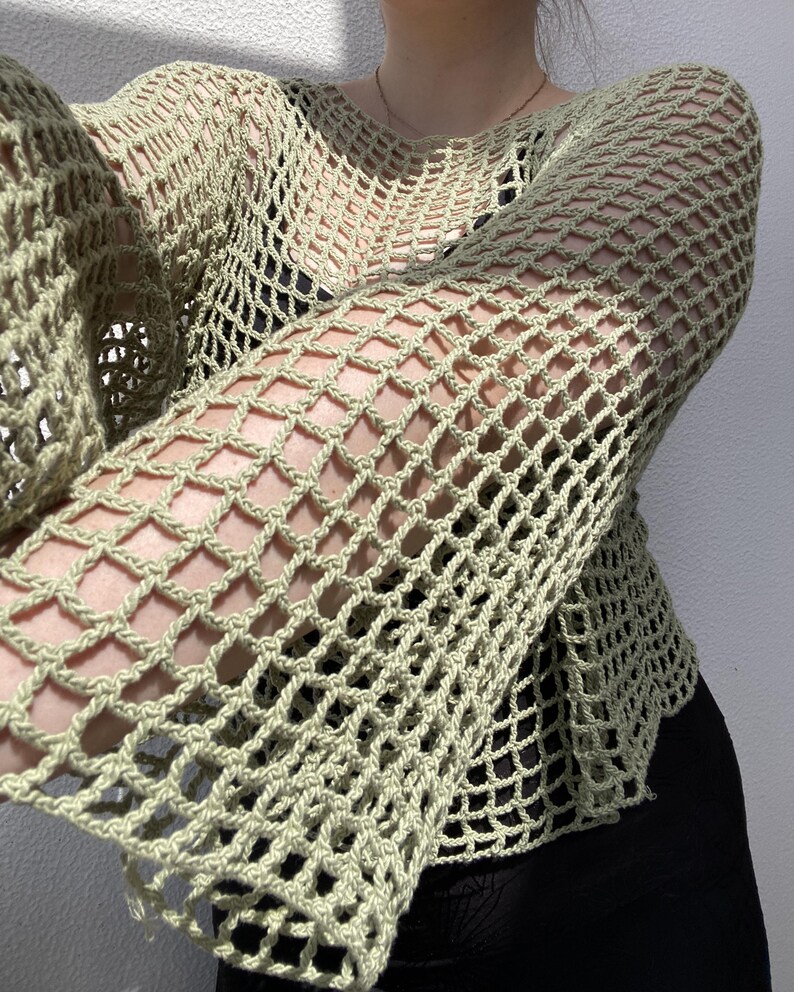 May include: A pale green, open-weave crochet top with long sleeves. The top has a loose, net-like pattern, creating a see-through effect. The garment is designed with a round neckline and a relaxed fit, suitable for layering or as a standalone piece.