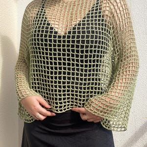 May include: A pale green, open-weave crochet top with long sleeves. The top is worn over a black garment, creating a layered effect. The crochet design features a grid-like pattern. The sleeves are flared at the wrist.