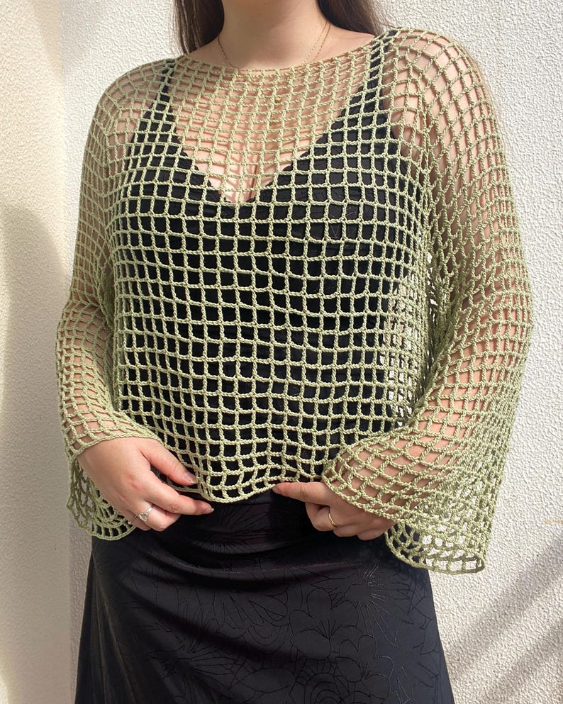 May include: A pale green, open-weave crochet top with long sleeves. The top is worn over a black garment, creating a layered effect. The crochet design features a grid-like pattern. The sleeves are flared at the wrist.