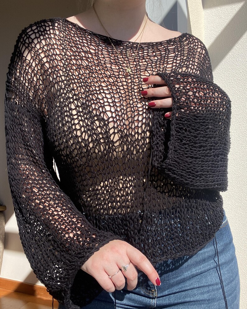 May include: Black open-weave knit top with long sleeves. The top has a boat neck and a loose, airy design. The sleeves are flared at the wrist. The top is paired with blue jeans.