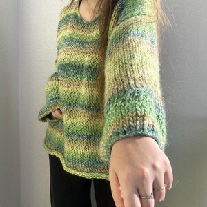 May include: A hand-knitted sweater in shades of green and yellow. The sweater has a scoop neck and long sleeves with ribbed cuffs. The yarn has a variegated color pattern, creating a striped effect. The sweater is a cozy, handmade garment.