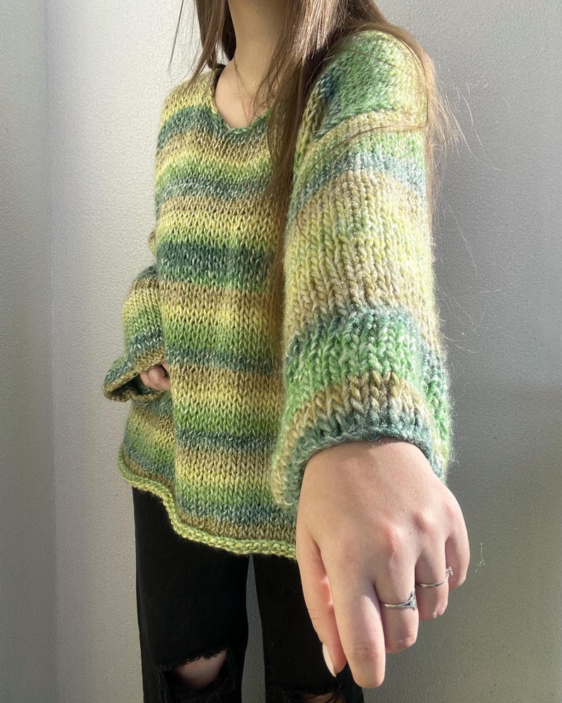May include: A hand-knitted sweater in shades of green and yellow. The sweater has a scoop neck and long sleeves with ribbed cuffs. The yarn has a variegated color pattern, creating a striped effect. The sweater is a cozy, handmade garment.