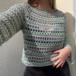 May include: A light green, white, and grey crochet jumper with long sleeves. The open weave design features a round neckline and a slightly cropped fit. The sleeves have a bell shape at the wrist. The jumper is handmade.