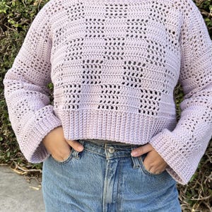 Pu&ograve; includere: Un maglione all'uncinetto lilla chiaro con scollo tondo e maniche lunghe. Il maglione presenta un motivo traforato e un orlo a coste. Le maniche sono svasate al polso. La persona indossa jeans blu.
