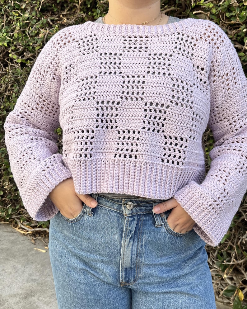 Pu&ograve; includere: Un maglione all'uncinetto lilla chiaro con scollo tondo e maniche lunghe. Il maglione presenta un motivo traforato e un orlo a coste. Le maniche sono svasate al polso. La persona indossa jeans blu.