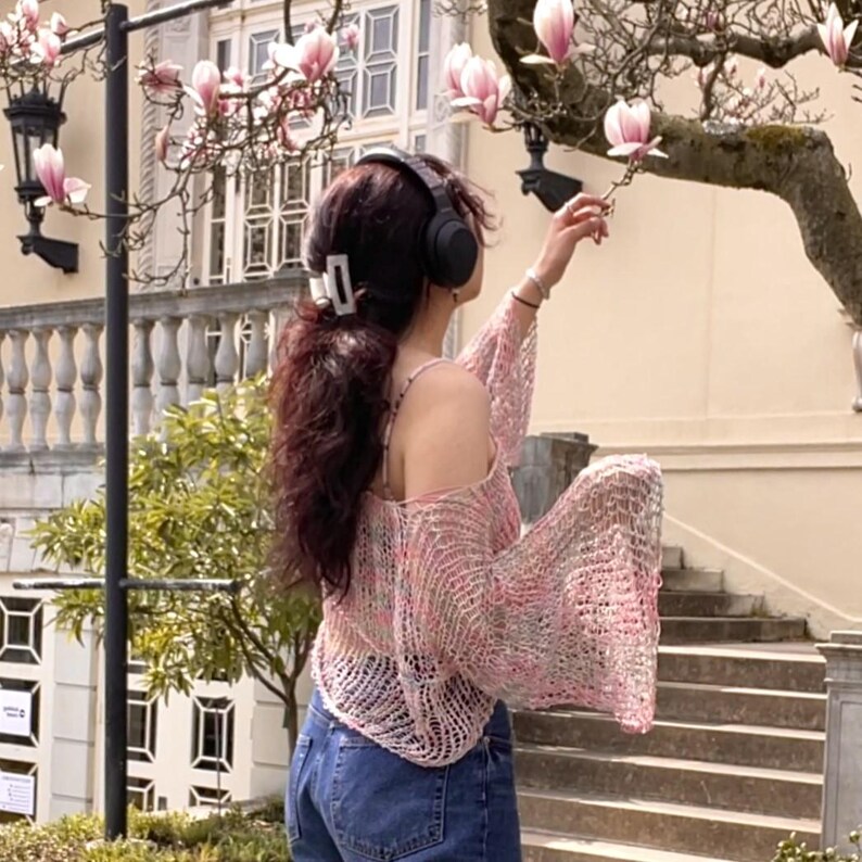 May include: A pink and white open-knit sweater with long sleeves and a scoop neck. The person is wearing blue jeans and black headphones. The background includes a flowering tree and a building with windows.