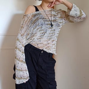 May include: Off-the-shoulder, open-knit sweater in cream with gold and brown stripes. The long-sleeved top has a loose, relaxed fit and a slightly cropped hem. The sweater is paired with black cargo pants.