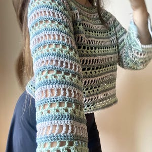 May include: A pastel-coloured crochet top with long sleeves. The top features a mix of light blue, green, and pale pink yarns, creating a striped pattern. The sleeves have an open, airy design. The garment is a cropped style.