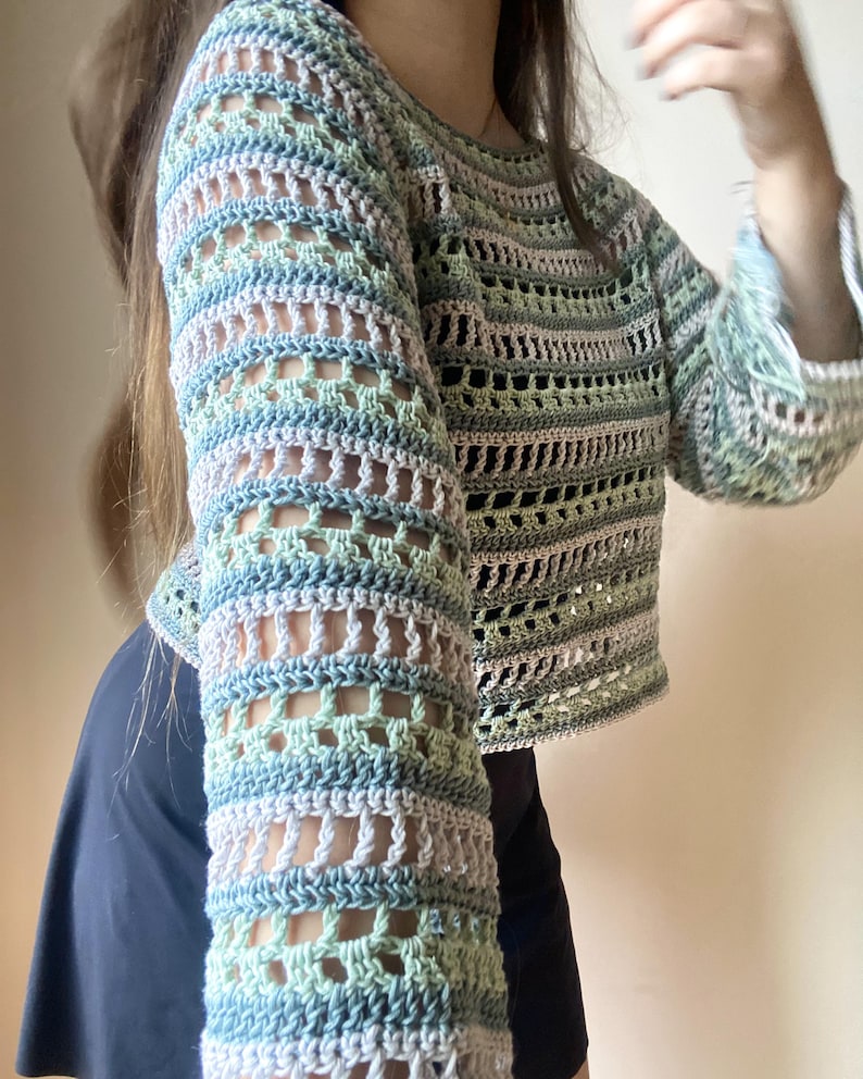 May include: A pastel-coloured crochet top with long sleeves. The top features a mix of light blue, green, and pale pink yarns, creating a striped pattern. The sleeves have an open, airy design. The garment is a cropped style.