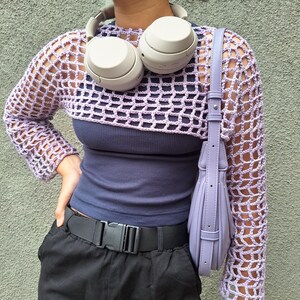 May include: A lilac crochet shrug with long sleeves is worn over a navy blue vest top. A matching lilac handbag hangs from the shoulder. A black belt with a buckle and black trousers are also visible. Grey headphones rest on the shoulders.