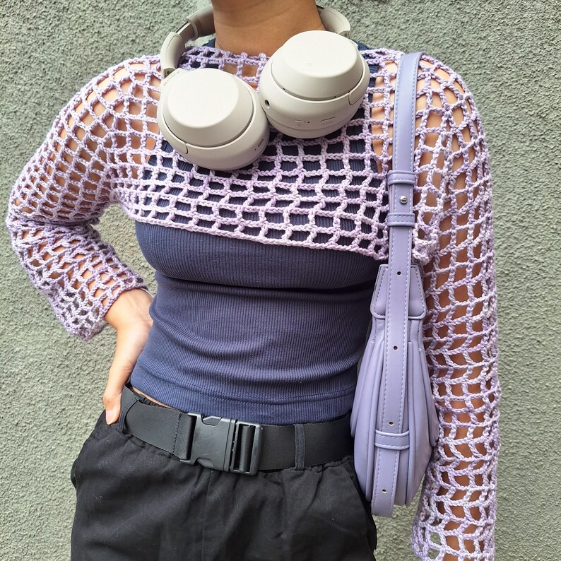 May include: A lilac crochet shrug with long sleeves is worn over a navy blue vest top. A matching lilac handbag hangs from the shoulder. A black belt with a buckle and black trousers are also visible. Grey headphones rest on the shoulders.
