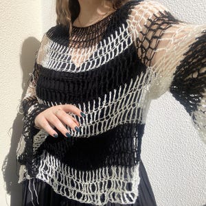 May include: A black and white open-weave sweater. The sweater features horizontal stripes of black and white, with long, flared sleeves. The garment is made of a loosely knit material, creating a see-through effect. The sweater is worn over a black top.