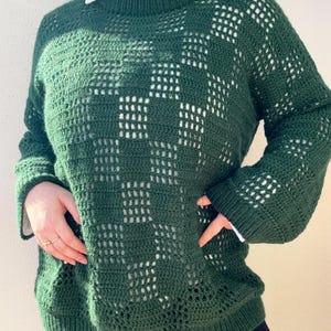 Pu&ograve; includere: Un maglione all'uncinetto verde scuro con un motivo a quadri. Il maglione ha maniche lunghe e girocollo. Sotto si vede una camicia bianca con colletto. Il maglione &egrave; realizzato con un filato morbido e strutturato.