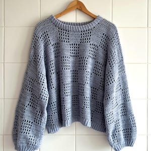 Pu&ograve; includere: Un maglione all'uncinetto azzurro con scollo tondo e maniche lunghe. Il maglione, realizzato in un morbido materiale a trama aperta, &egrave; appeso a una gruccia di legno contro un muro piastrellato bianco. Le maniche sono leggermente a sbuffo.