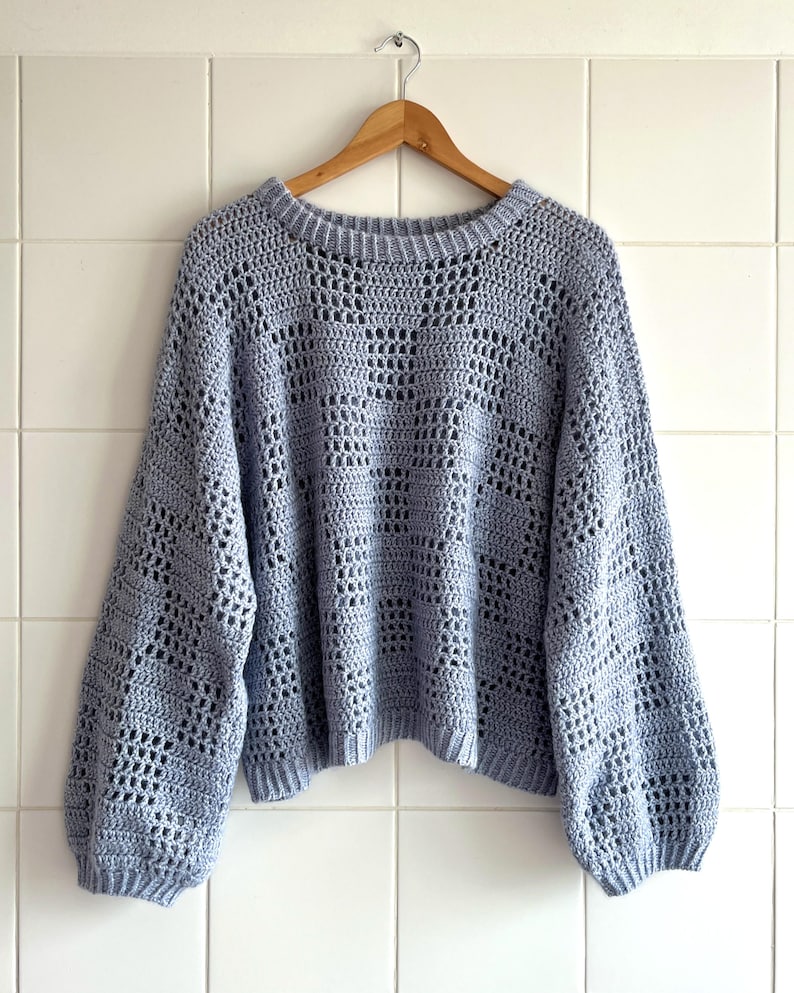 Pu&ograve; includere: Un maglione all'uncinetto azzurro con scollo tondo e maniche lunghe. Il maglione, realizzato in un morbido materiale a trama aperta, &egrave; appeso a una gruccia di legno contro un muro piastrellato bianco. Le maniche sono leggermente a sbuffo.