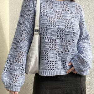 Pu&ograve; includere: Un maglione all'uncinetto azzurro con scollo tondo e maniche lunghe. Il maglione ha un design aperto e arioso. Una borsa a tracolla bianca con fibbia argentata &egrave; appesa a sinistra. La persona indossa una gonna nera.