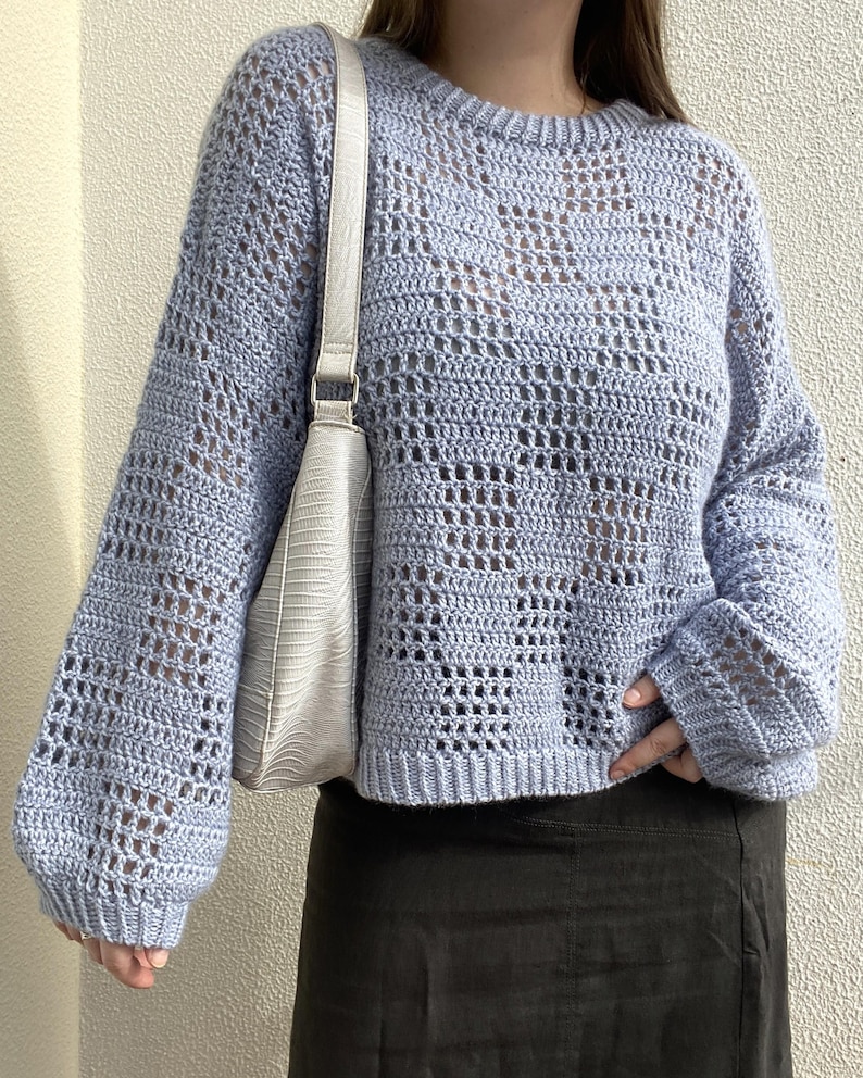 Pu&ograve; includere: Un maglione all'uncinetto azzurro con scollo tondo e maniche lunghe. Il maglione ha un design aperto e arioso. Una borsa a tracolla bianca con fibbia argentata &egrave; appesa a sinistra. La persona indossa una gonna nera.