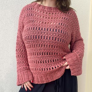 May include: A dusty rose crochet sweater with a round neckline and long sleeves. The sweater features a textured openwork design throughout the body and sleeves, with ribbed cuffs and hem. The garment is made of a soft, possibly wool or cotton yarn.