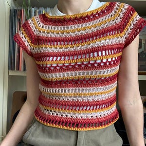 May include: A handmade crochet top in a striped pattern of red, yellow, and cream colours. The short-sleeved top has a round neckline and a slightly open weave design. The top is worn over a white undershirt.
