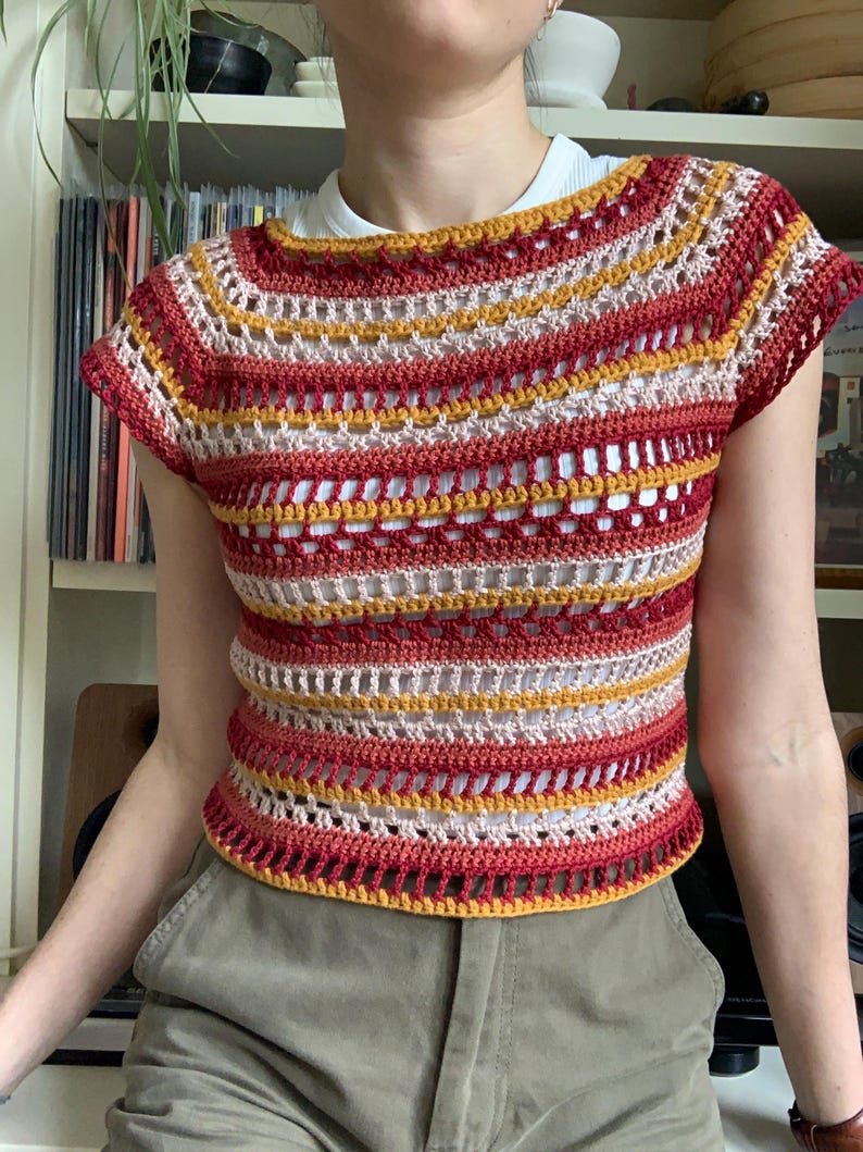 May include: A handmade crochet top in a striped pattern of red, yellow, and cream colours. The short-sleeved top has a round neckline and a slightly open weave design. The top is worn over a white undershirt.