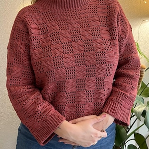 Pu&ograve; includere: Un maglione all'uncinetto color rosso mattone con collo alto e maniche lunghe. Il maglione presenta un motivo geometrico aperto sul corpo e sulle maniche. Le maniche hanno un polsino a coste. Il maglione &egrave; abbinato a jeans blu.