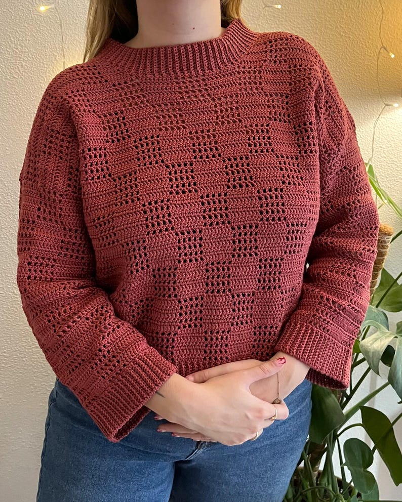 Pu&ograve; includere: Un maglione all'uncinetto color rosso mattone con collo alto e maniche lunghe. Il maglione presenta un motivo geometrico aperto sul corpo e sulle maniche. Le maniche hanno un polsino a coste. Il maglione &egrave; abbinato a jeans blu.
