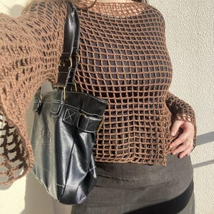 May include: A brown, open-weave crochet top with long sleeves. A black leather handbag with white stitching and a logo is hanging from the shoulder. The outfit is completed with a dark skirt.