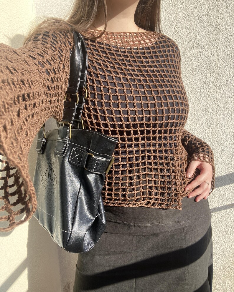 May include: A brown, open-weave crochet top with long sleeves. A black leather handbag with white stitching and a logo is hanging from the shoulder. The outfit is completed with a dark skirt.