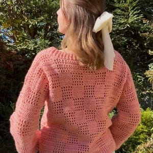 Pu&ograve; includere: Un maglione all'uncinetto color corallo chiaro con scollo tondo e maniche lunghe. Il maglione presenta un design a trama aperta strutturata. Un accessorio per capelli bianco &egrave; visibile sullo sfondo.