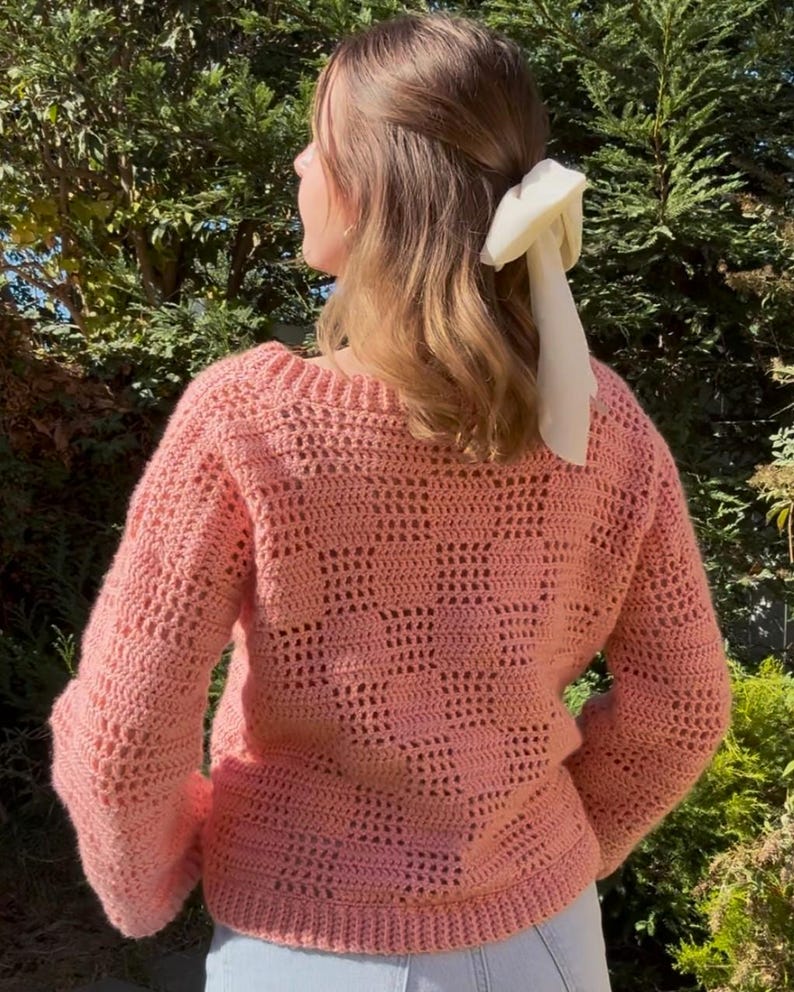 Pu&ograve; includere: Un maglione all'uncinetto color corallo chiaro con scollo tondo e maniche lunghe. Il maglione presenta un design a trama aperta strutturata. Un accessorio per capelli bianco &egrave; visibile sullo sfondo.