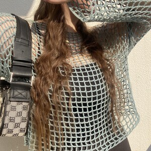 May include: A pale blue crochet top with a square net design and long sleeves. The top is worn over a black garment. A black handbag with a black strap and a grey and black pattern is visible on the left.
