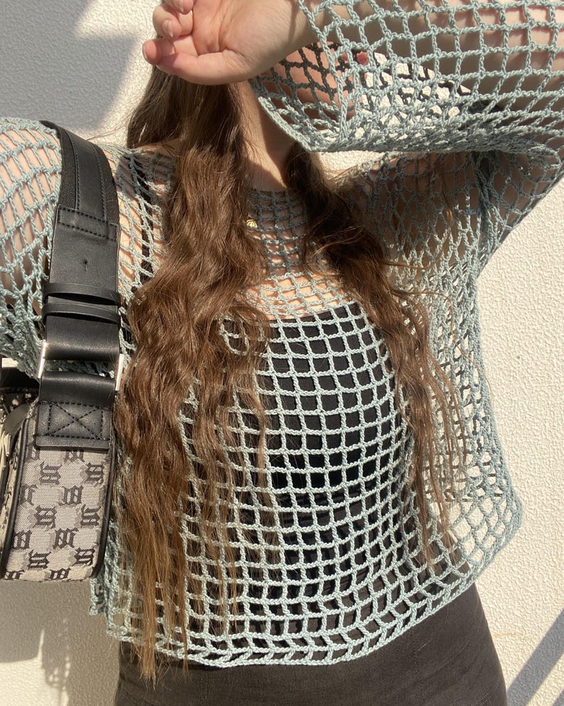 May include: A pale blue crochet top with a square net design and long sleeves. The top is worn over a black garment. A black handbag with a black strap and a grey and black pattern is visible on the left.