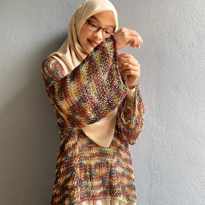 May include: A multi-colored knit top with bell sleeves. The top features a mix of red, yellow, green, and brown threads. The top is paired with a beige dress and a matching head covering. The background is a solid gray wall.