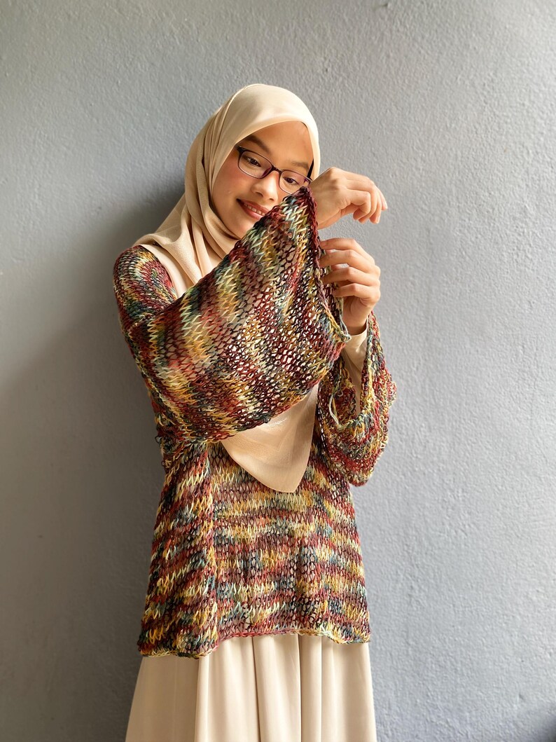 May include: A multi-colored knit top with bell sleeves. The top features a mix of red, yellow, green, and brown threads. The top is paired with a beige dress and a matching head covering. The background is a solid gray wall.