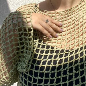 May include: A light green, open-weave crochet top. The top is worn over a black garment, creating a contrast. The crochet design features a grid-like pattern. The sleeves are long and the neckline is round. A silver ring is visible on the hand.