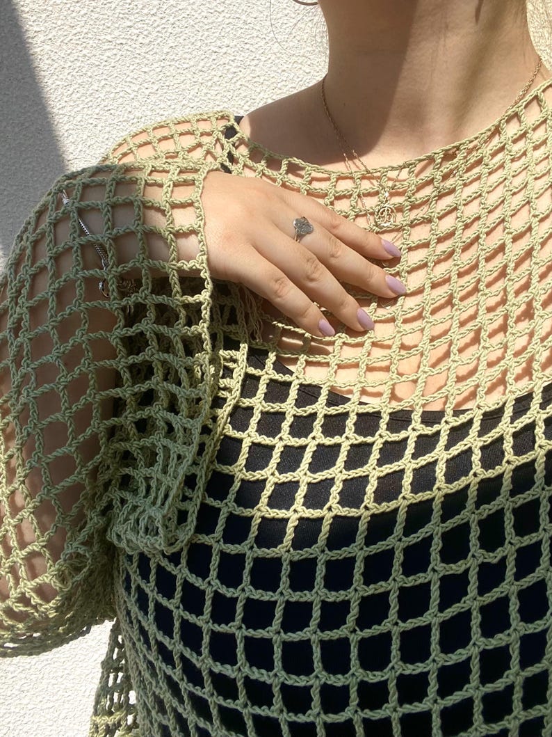 May include: A light green, open-weave crochet top. The top is worn over a black garment, creating a contrast. The crochet design features a grid-like pattern. The sleeves are long and the neckline is round. A silver ring is visible on the hand.