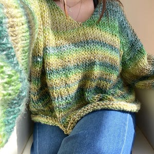 May include: A hand-knitted, oversized V-neck sweater in shades of green, yellow, and blue. The sweater features a striped pattern and a relaxed fit. The sleeves are long and the knit is chunky, giving it a cozy appearance. The sweater is paired with blue jeans.