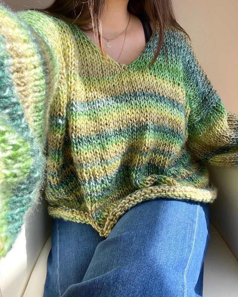 May include: A hand-knitted, oversized V-neck sweater in shades of green, yellow, and blue. The sweater features a striped pattern and a relaxed fit. The sleeves are long and the knit is chunky, giving it a cozy appearance. The sweater is paired with blue jeans.