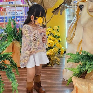 May include: A young child wearing a pastel-colored knit sweater, a white skirt, brown boots, and tan socks. The child is pointing at something. The background includes yellow flowers, decorative elements, and a sign.