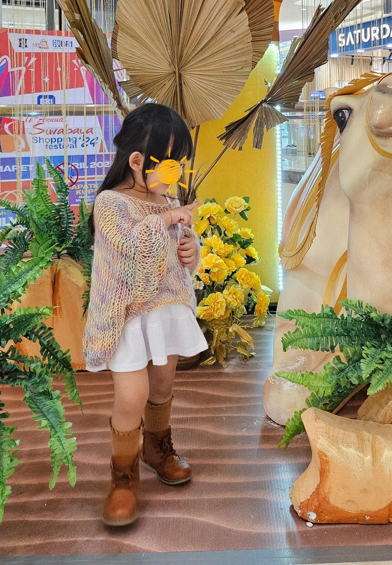 May include: A young child wearing a pastel-colored knit sweater, a white skirt, brown boots, and tan socks. The child is pointing at something. The background includes yellow flowers, decorative elements, and a sign.